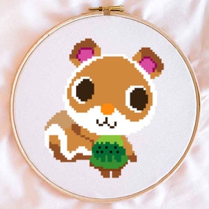 Puede incluir: Un bastidor de bordado de punto de cruz con un diseño de hámster pixelado. El hámster tiene pelaje marrón, ojos negros grandes y un atuendo verde. El bordado está sobre un fondo de tela blanca, enmarcado por un aro de madera.