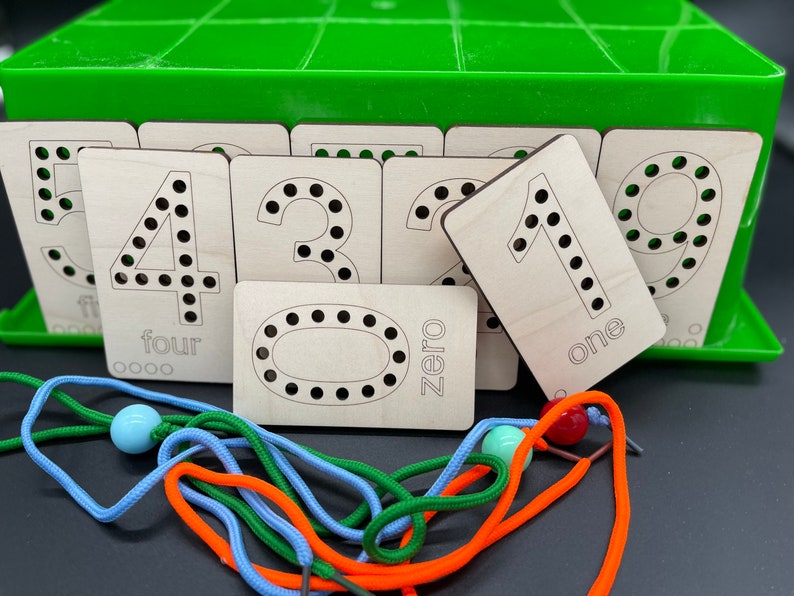 May include: Wooden number tiles with holes for lacing, featuring the numbers 0 through 5. The tiles are arranged in a green plastic storage box.  The tiles are labeled with the number and the word for the number.  There are also colorful laces and beads.
