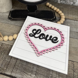 Pu&ograve; includere: Un cartello di legno bianco con una cornice a forma di cuore rosa e la parola "Love" in lettere nere.