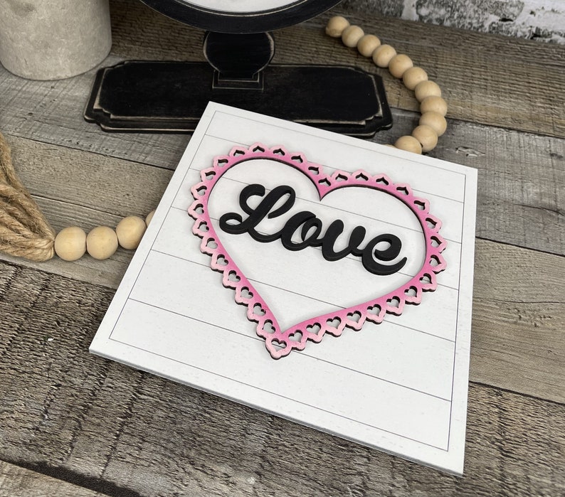 Pu&ograve; includere: Un cartello di legno bianco con una cornice a forma di cuore rosa e la parola "Love" in lettere nere.