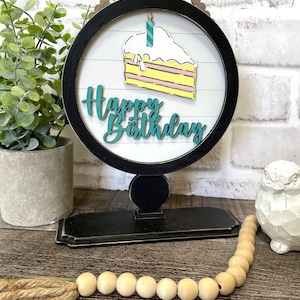 May include: A black and white wooden birthday sign with a slice of cake and a teal candle. The words "Happy Birthday" are in teal script. The sign is on a black stand with a wooden bead garland.