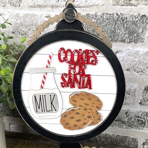 May include: A round wooden sign with a black frame and white woodgrain background. The sign says "Cookies for Santa" in red letters with white polka dots. There is a milk bottle with a red and white striped straw and a stack of chocolate chip cookies.
