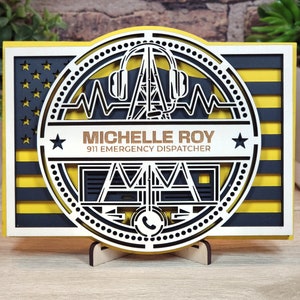 May include: A wooden plaque with a yellow and black American flag design. The center features a circle with a stylized image of a 911 dispatcher's console, headphones, and a heartbeat monitor. The text "Michelle Roy 911 Emergency Dispatcher" is displayed in gold lettering.