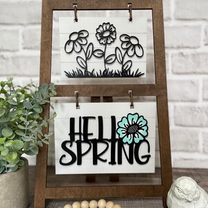 May include: A wooden easel with two signs. The top sign has a black silhouette of flowers. The bottom sign says "Hello Spring" with a teal flower.