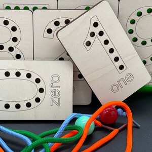 May include: Wooden number cards with holes for lacing, featuring the numbers zero through nine. The cards are light brown and have black outlines around the numbers. The word "zero" is written on the card with the number zero. The word "one" is written on the card with the number one.  The cards are arranged in a pile with the number one card on top.  There are colorful shoelaces and beads in the foreground.