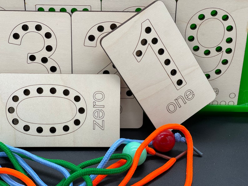 May include: Wooden number cards with holes for lacing, featuring the numbers zero through nine. The cards are light brown and have black outlines around the numbers. The word "zero" is written on the card with the number zero. The word "one" is written on the card with the number one.  The cards are arranged in a pile with the number one card on top.  There are colorful shoelaces and beads in the foreground.