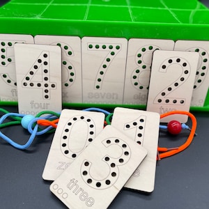 May include: Wooden number cards with holes for threading. The cards are arranged in a green plastic box. The numbers are 0, 1, 2, 3, 4, 5, 6, 7, 8, and 9. The cards are labeled with the number and the word for the number.  The cards are used for teaching children to count.