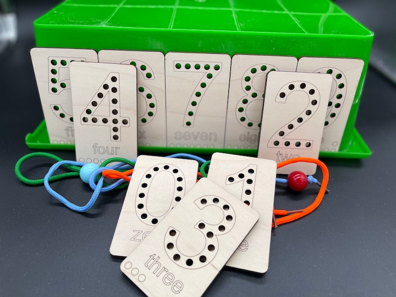 May include: Wooden number cards with holes for threading. The cards are arranged in a green plastic box. The numbers are 0, 1, 2, 3, 4, 5, 6, 7, 8, and 9. The cards are labeled with the number and the word for the number.  The cards are used for teaching children to count.