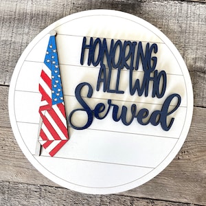 May include: A white wooden round sign with a blue and red American flag on the left side and the text "Honoring All Who Served" in blue lettering.