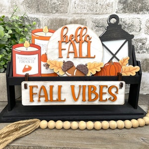 May include: A black tiered shelf with fall-themed decorations. The shelf features a round wooden sign that says "hello fall", two candles with fall-themed labels, a lantern, a pumpkin, and a sign that says "fall vibes".