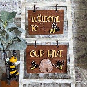 May include: Welcome to Our Hive sign with two wooden bees and a honeycomb. The sign is on a white wooden easel with a distressed finish.