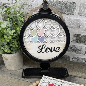 May include: A black wooden stand with a round, white sign featuring a cutout design of hearts. The sign says "Love" in black cursive lettering. There are five smaller hearts in the center of the sign, painted in shades of pink, blue, and green.