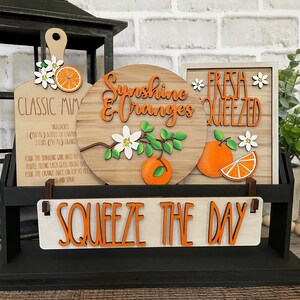 May include: A wooden sign with the text "Squeeze the Day" in orange letters. There are three smaller signs above it, one with a recipe for "Classic Mimosa", one with the text "Sunshine & Oranges" and a branch of orange blossoms, and one with the text "Fresh Squeezed" and a slice of orange.