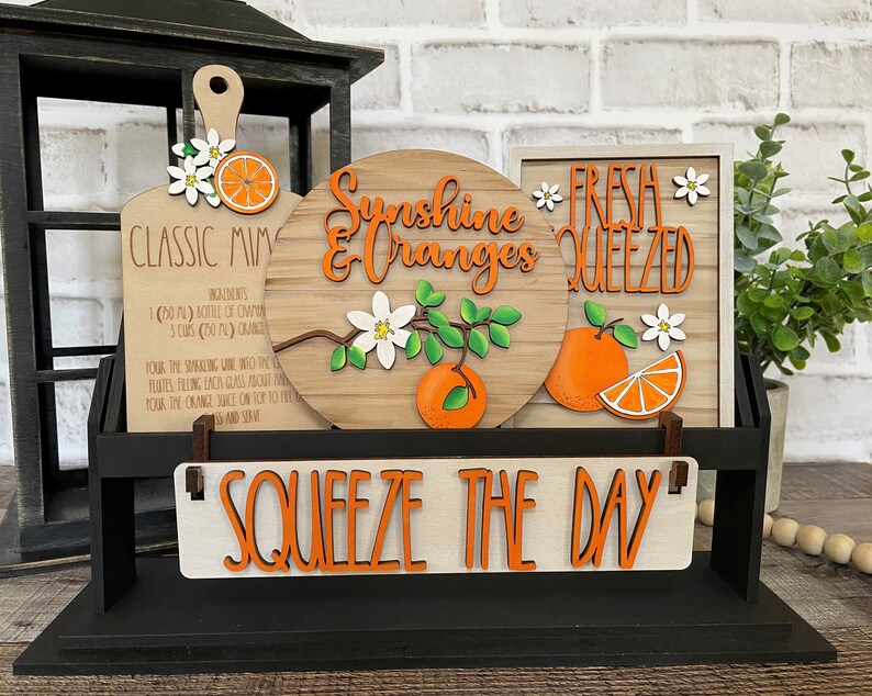 May include: A wooden sign with the text "Squeeze the Day" in orange letters. There are three smaller signs above it, one with a recipe for "Classic Mimosa", one with the text "Sunshine & Oranges" and a branch of orange blossoms, and one with the text "Fresh Squeezed" and a slice of orange.