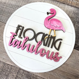 May include: A white round wooden sign with the words "Flocking Fabulous" in black and pink letters. A pink flamingo with black eyes is standing on the sign.
