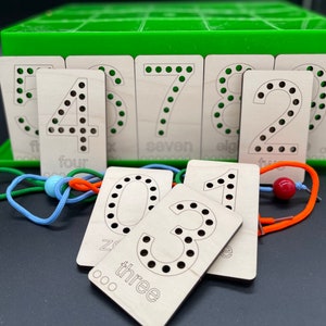 May include: Wooden number cards with holes for lacing. The cards feature the numbers 1 through 8, with the words "one" through "eight" written below each number. The cards are arranged in a green box with a lid.