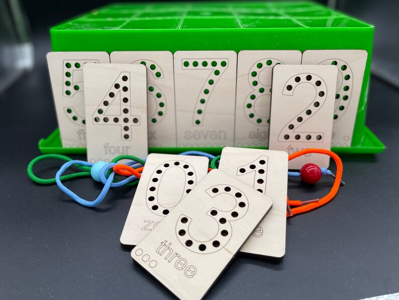May include: Wooden number cards with holes for lacing. The cards feature the numbers 1 through 8, with the words "one" through "eight" written below each number. The cards are arranged in a green box with a lid.