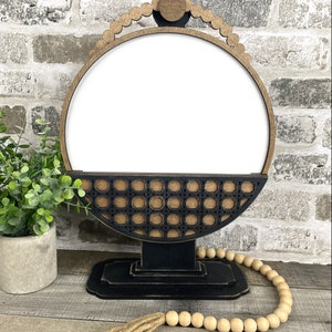 May include: A black and brown wooden decorative stand with a round, white center. The stand has a woven pattern and a beaded trim. A wooden bead garland with a jute tassel is in the foreground.