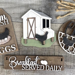 May include: Four wooden signs with a farmhouse theme. The signs feature a chicken coop, a chicken in a nest, a chicken coop with the text "The Chick Inn", and a sign that says "Breakfast Served Daily".