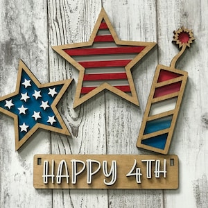 May include: Three wooden cutouts in red, white, and blue, featuring a star with stripes, a star with white stars, and a firecracker. The cutouts are arranged on a white wood background with a wooden sign that reads "HAPPY 4TH".