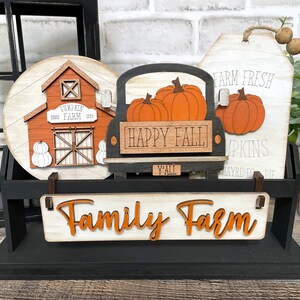 May include: A wooden tiered stand with a black base and white shelves. The stand features a round wooden sign with a red barn and the text "Pumpkin Farm Since 1923", a black truck with the text "Happy Fall Y'all" and three orange pumpkins, and a white sign with the text "Farm Fresh Pumpkins Hayrides Pie".