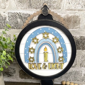 May include: A round wooden sign with a blue and white rainbow design. The rainbow has gold stars and a candle in the center. The sign says "Love & Light".