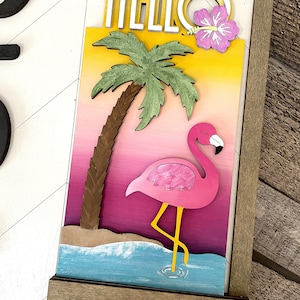 May include: A wooden sign with the word "HELLO" in white letters with a yellow outline. The sign features a pink flamingo standing in blue water with a palm tree and a pink hibiscus flower.