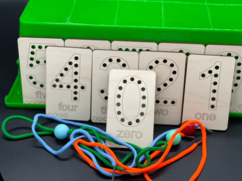 May include: Wooden number cards with black dots and the numbers 0 through 5 written on them. The cards are arranged in a row with the number 0 on the left and the number 5 on the right. There are also colorful laces and beads on the table.