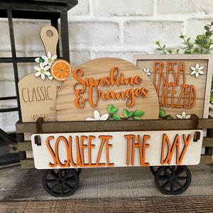 May include: A wooden wagon with a white sign that says "Squeeze the Day" in orange letters. The wagon is decorated with wooden signs that say "Sunshine & Oranges", "Classic Mimosas", and "Fresh Squeezed". The signs are decorated with orange slices and white flowers.