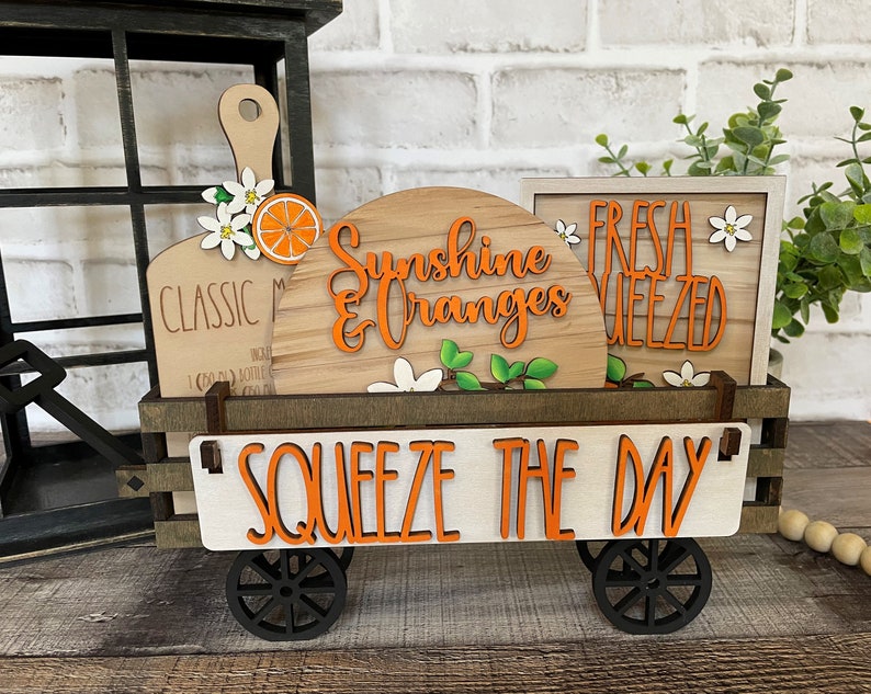 May include: A wooden wagon with a white sign that says "Squeeze the Day" in orange letters. The wagon is decorated with wooden signs that say "Sunshine & Oranges", "Classic Mimosas", and "Fresh Squeezed". The signs are decorated with orange slices and white flowers.