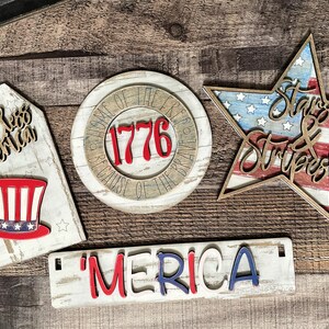 May include: Four wooden signs with a patriotic theme. The signs feature the words "God Bless America", "1776", "Stars & Stripes", and "'MERICA". The signs are painted in red, white, and blue.