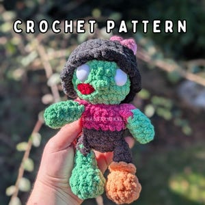 May include: A crochet pattern for a plush doll featuring a green face, white eyes, a red mouth, and a dark grey hat. The doll wears a pink shirt, dark grey trousers, and has one green leg and one orange leg. The text CROCHET PATTERN is at the top.