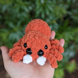 Puede incluir: Un juguete de peluche de ara&ntilde;a de ganchillo naranja y blanco con ojos negros. La ara&ntilde;a est&aacute; sentada en la palma de una mano.