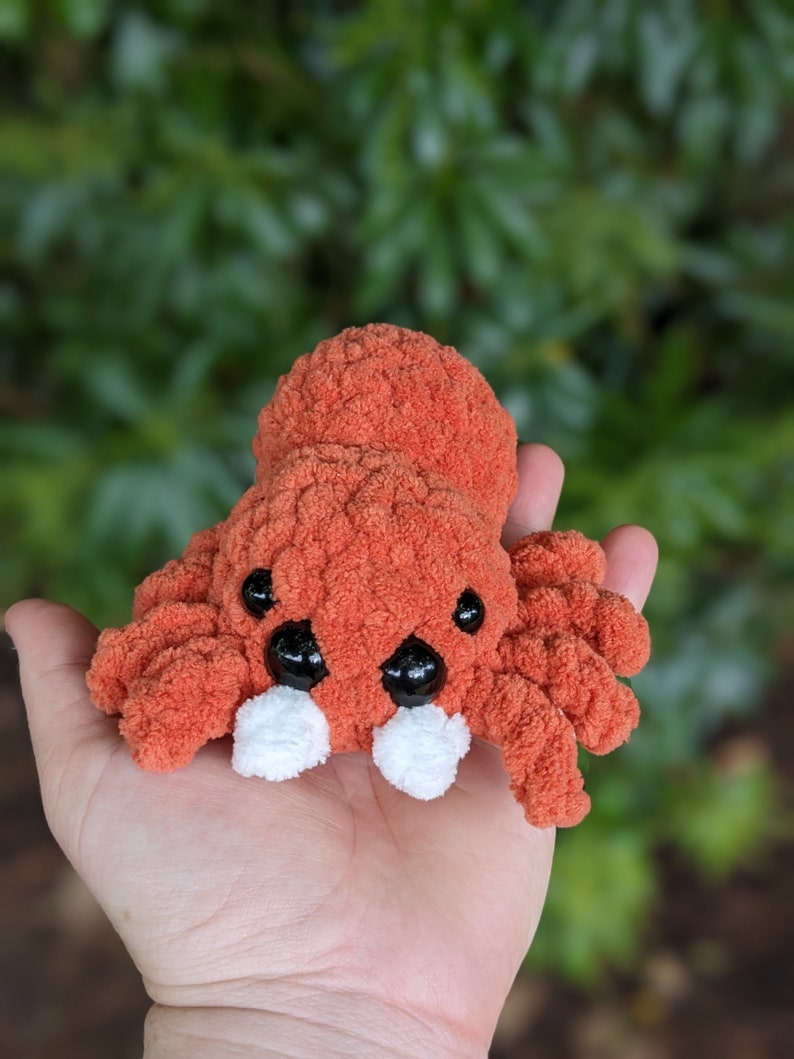 Puede incluir: Un juguete de peluche de ara&ntilde;a de ganchillo naranja y blanco con ojos negros. La ara&ntilde;a est&aacute; sentada en la palma de una mano.