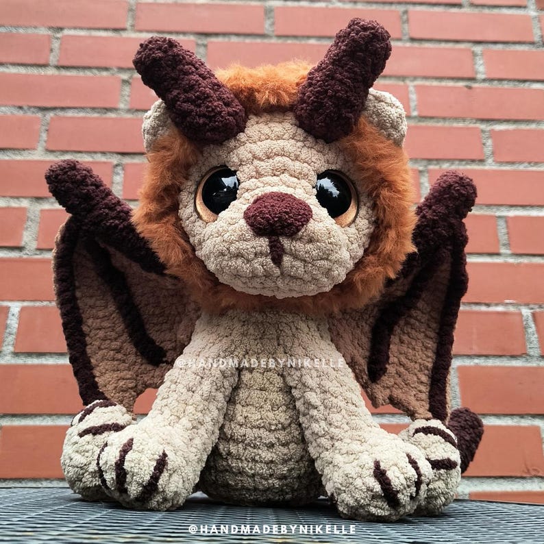 Op de afbeelding: Een handgemaakt gehaakt knuffel in de vorm van een mythisch wezen. Het speelgoed heeft een leeuwachtig hoofd met bruine hoorns, grote zwarte ogen en bruine vleugels. Het lichaam is beige en bruin, met een zacht, getextureerd uiterlijk. Het speelgoed zit op een metalen oppervlak.
