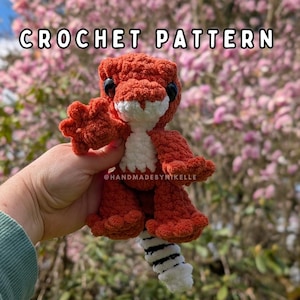 Può includere: Un dinosauro all'uncinetto fatto a mano nei toni dell'arancione e del bianco, con occhi neri. Il testo "CROCHET PATTERN" è visualizzato nella parte superiore dell'immagine. Il dinosauro ha una coda a strisce ed è tenuto in mano.