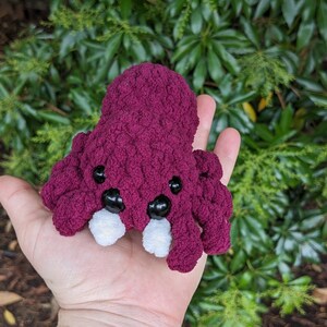 Puede incluir: Un peque&ntilde;o juguete de peluche de pulpo tejido a crochet con un cuerpo burdeos, dientes blancos y ojos negros. El pulpo est&aacute; en la mano de una persona.
