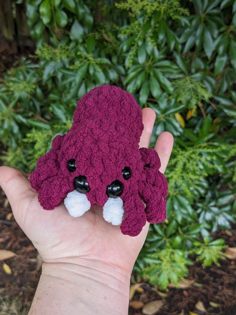 Puede incluir: Un peque&ntilde;o juguete de peluche de pulpo tejido a crochet con un cuerpo burdeos, dientes blancos y ojos negros. El pulpo est&aacute; en la mano de una persona.