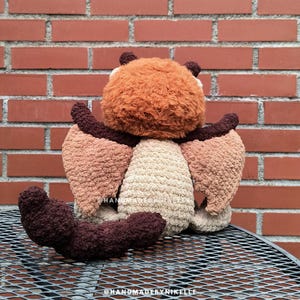 Op de afbeelding: Een handgemaakt gehaakt knuffel in de vorm van een vleermuis, van achteren gezien. De vleermuis heeft een bruin, beige en bruin kleurenschema. De knuffel heeft een pluizige kop, getextureerde vleugels en een gekrulde staart.