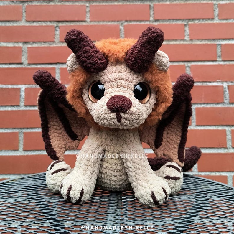 Op de afbeelding: Een handgemaakte gehaakte draak knuffel in bruin, beige en oranje tinten. De draak heeft grote, expressieve ogen, hoorns en vleugels. De knuffel zit op een metalen oppervlak voor een bakstenen muur. De tekst "@HANDMADEBYNIKELLE" is zichtbaar.