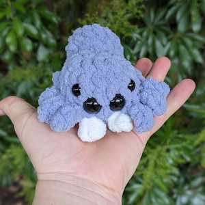 Puede incluir: Un peque&ntilde;o juguete de peluche tejido a crochet en forma de pulpo morado con ojos blancos y pies blancos. El pulpo est&aacute; sentado en la palma de una mano.
