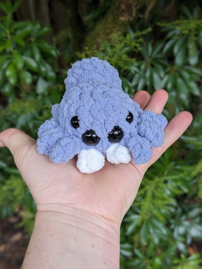 Puede incluir: Un peque&ntilde;o juguete de peluche tejido a crochet en forma de pulpo morado con ojos blancos y pies blancos. El pulpo est&aacute; sentado en la palma de una mano.