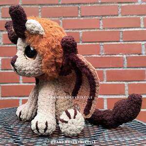 Op de afbeelding: Een handgemaakte gehaakte draak knuffel in bruin, beige en oranje tinten. De draak heeft grote ogen, hoorns en vleugels. De knuffel zit op een metalen oppervlak voor een bakstenen muur. De tekst "handmadebynikelle" is zichtbaar.