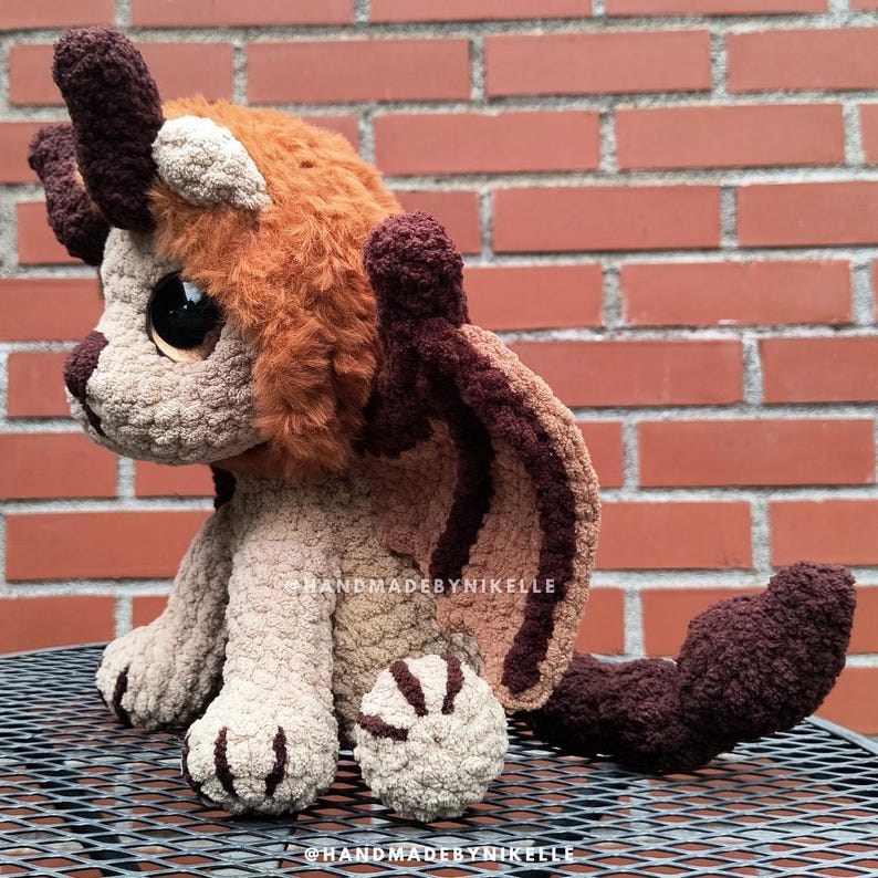 Op de afbeelding: Een handgemaakte gehaakte draak knuffel in bruin, beige en oranje tinten. De draak heeft grote ogen, hoorns en vleugels. De knuffel zit op een metalen oppervlak voor een bakstenen muur. De tekst "handmadebynikelle" is zichtbaar.