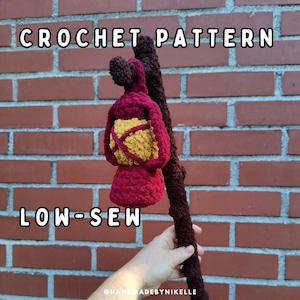 May include: A burgundy and yellow crochet lantern attached to a dark brown crocheted branch. The text "CROCHET PATTERN" and "LOW-SEW" are displayed in white block letters. The lantern has a textured appearance.