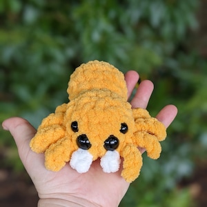 Puede incluir: Una ara&ntilde;a naranja de crochet con ojos negros y colmillos blancos. La ara&ntilde;a est&aacute; sentada en la palma de una mano.