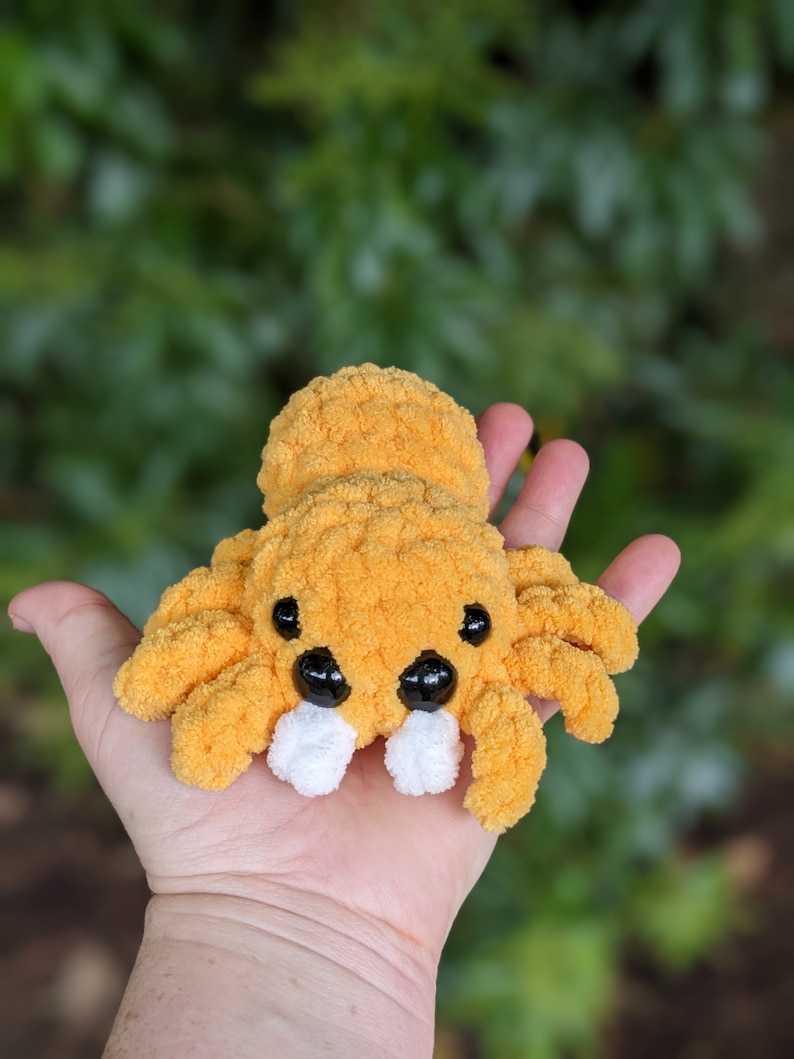 Puede incluir: Una ara&ntilde;a naranja de crochet con ojos negros y colmillos blancos. La ara&ntilde;a est&aacute; sentada en la palma de una mano.