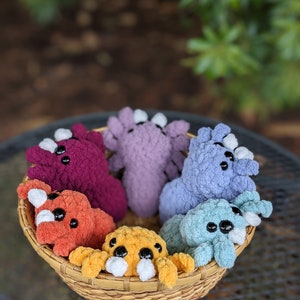 Puede incluir: Una cesta llena de seis juguetes de peluche tejidos a crochet en varios colores, incluyendo morado, azul, naranja y amarillo. Los juguetes tienen ojos grandes y tienen forma de peque&ntilde;as criaturas.