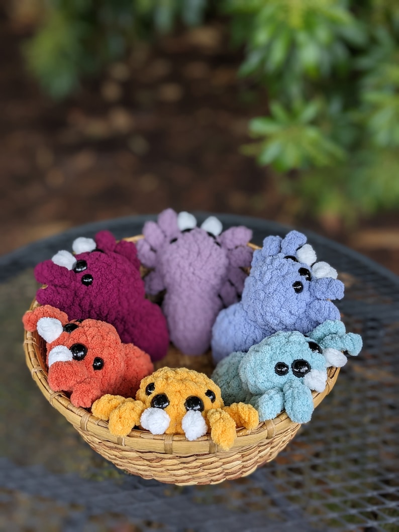 Puede incluir: Una cesta llena de seis juguetes de peluche tejidos a crochet en varios colores, incluyendo morado, azul, naranja y amarillo. Los juguetes tienen ojos grandes y tienen forma de peque&ntilde;as criaturas.