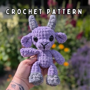 May include: A handmade crochet plush toy shaped like a goat, featuring light purple yarn for the body and dark gray yarn for the horns and hooves. The toy has large black plastic safety eyes and a stitched nose. The text CROCHET PATTERN is visible.